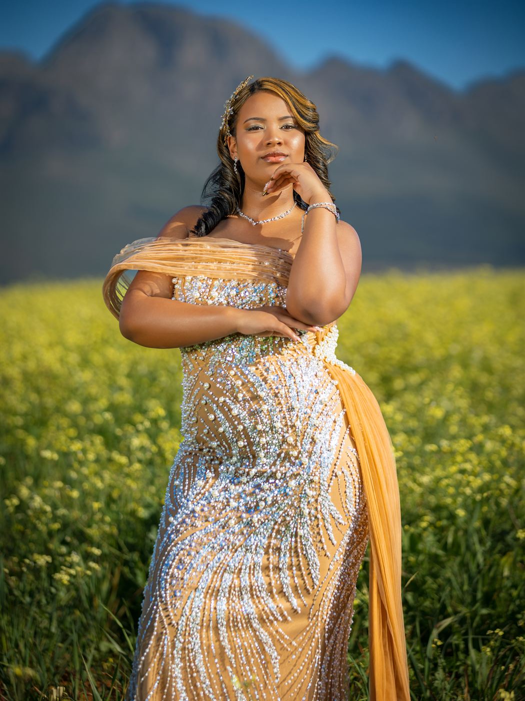 Young woman in a beaded gown standing in a canola field below the mountains