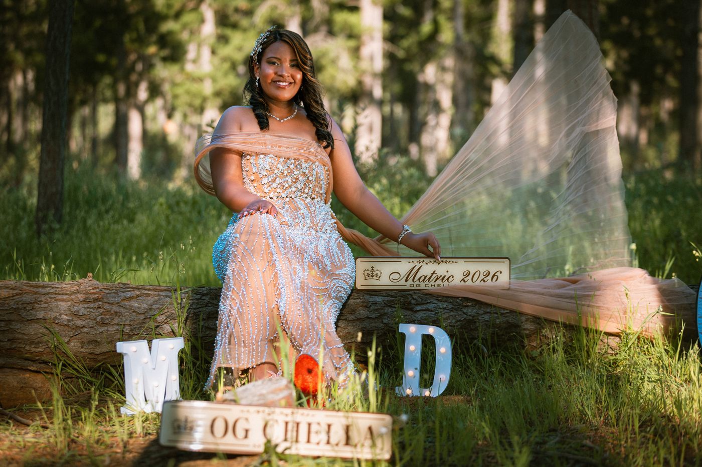 Young woman in a beaded gown seated on a log beside a Matric 2026 sign