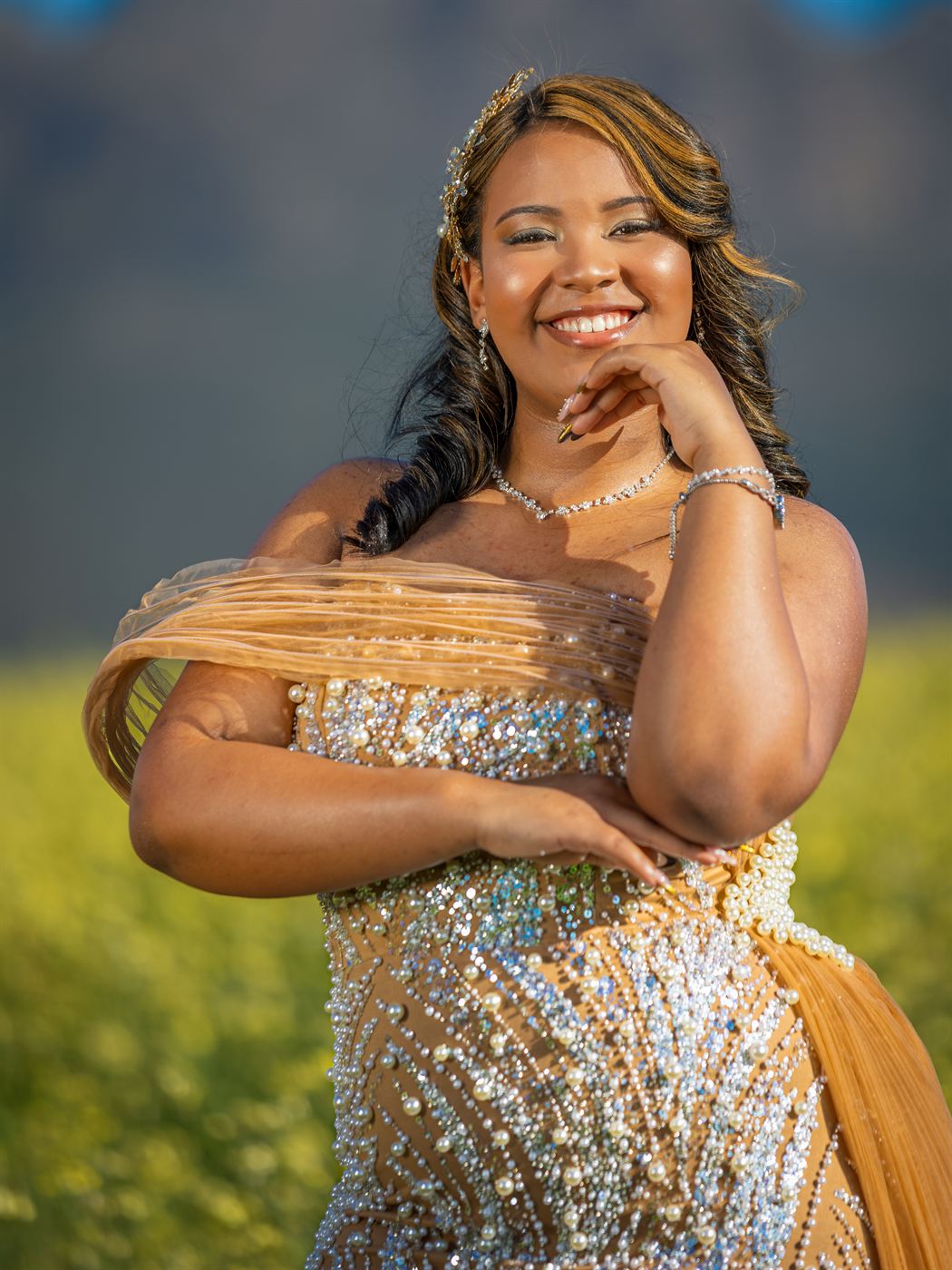 Young woman in a beaded gown smiling, hand resting at her chin, in a canola field